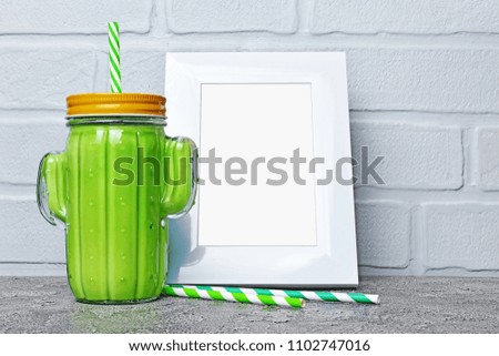 Glass jar of detox helthy green smoothy in the shape of cactus and mock-up of white frame with copy space for poster on gray concrete and brick wall background
