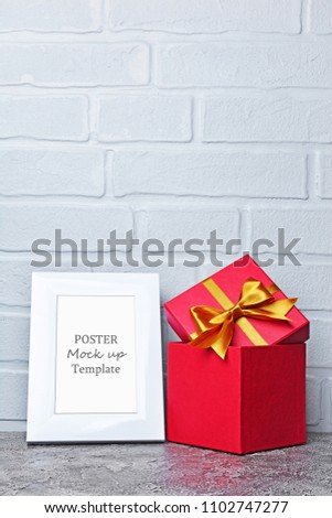 Mock-up of white frame with copy space for poster and red classic gift box with golden satin bow on gray concrete and brick wall background