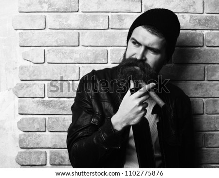 Bearded man, long beard. Brutal caucasian serious unshaven hipster smoking cigar with black leather jacket, hat and white shirt on beige brick wall studio background