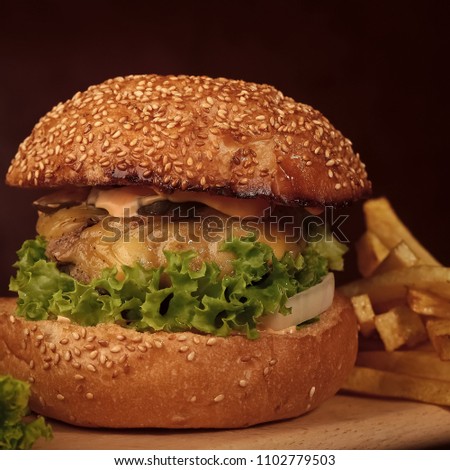 Tasty appetizing fresh burger of green lettuce cheese bacon slice meat cutlet and white bread bun with sesame seeds and potato chips on wooden table and potato chips, square picture