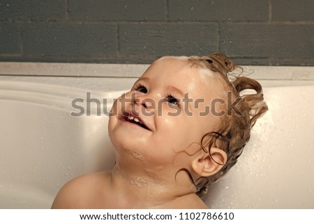 Kids playing with toys. Small happy pretty baby boy sitting in white bathroom with wet foam hair looking away, horizontal picture