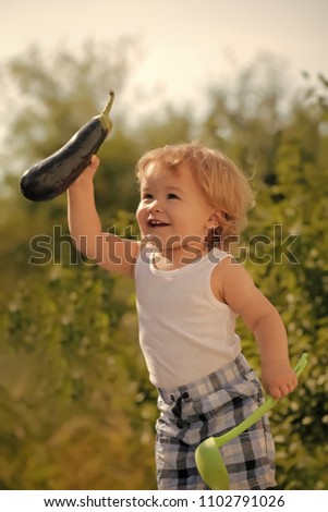 Kid play. Happy little smiling boy in checkered summer shorts at picnic with ladle and purple eggplant standing outdoor on green natural background sunny day, vertical picture