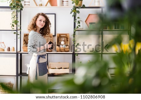 View through blurry leaves on a female employee in apron holding an organic skincare product in a shop with vegan cosmetics