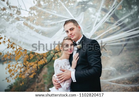 A young groom in a black suit and a beautiful bride in a lace dress embrace under a veil against the background of a tree with yellow leaves. Autumn wedding portrait of cute newlyweds. Film effect.