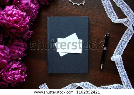 Flat lay home office desk. Female workspace with clear business card, pink peonies bouquet, silver accessories, black notebook on dark background. Top view feminine background.