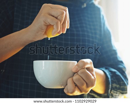 Female hand squeezing a lemon half into a white cup on blurred background of her blue plaid or tartan blouse. Food and drink ingredients preparing or bakery processing concept. (selective focus)