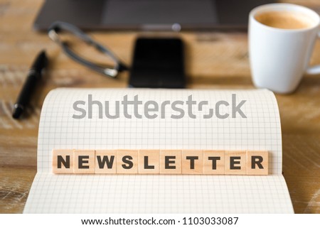 Closeup on notebook over wood table background, focus on wooden blocks with letters making Newsletter text. Concept image. Laptop, glasses, pen and mobile phone in defocused background