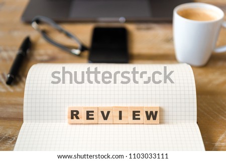 Closeup on notebook over wood table background, focus on wooden blocks with letters making Review text. Concept image. Laptop, glasses, pen and mobile phone in defocused background Royalty-Free Stock Photo #1103033111