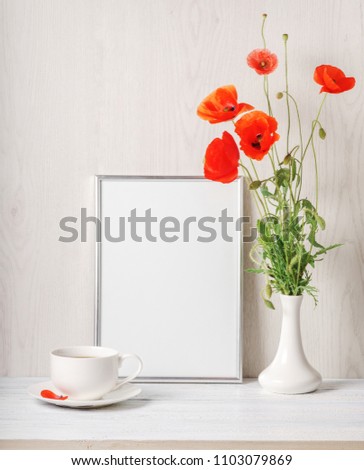 Red poppies bouquet in vase, tee cup and frame for text near wall on background.