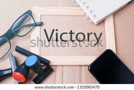 Conceptual image of motivational word of "VICTORY" with a multi colour stamp, a spectacle, smartphone and a note book over clean board background