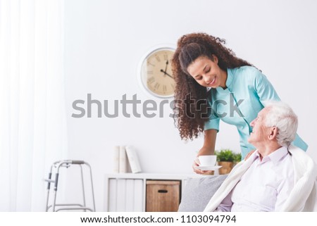 Young caregiver serving a cup of tea to a happy, older man in a retirement home