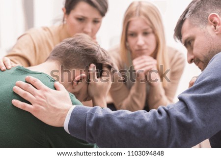Close-up of a devastated young man holding his head in his hands and friends supporting him during group therapy Royalty-Free Stock Photo #1103094848