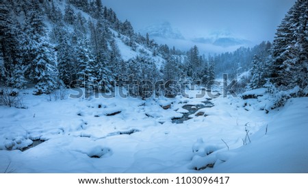 Snowscape river flowing towards fog-covered mountains in the background. Royalty-Free Stock Photo #1103096417