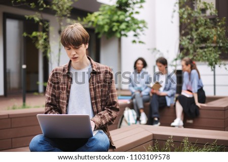 Photo of upset young man sitting on bench and sadly working on laptop while spending time in courtyard of university with students on background