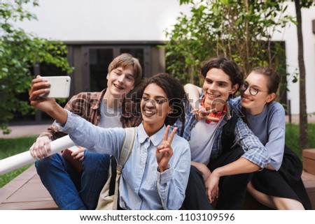 Group of young smiling students sitting and taking cute photos on cellphone while spending time together in courtyard of university 
