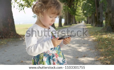 CLOSE UP: Adorable baby girl watching fun cartoons on her mother's cell phone during family walk down tree promenade. Lovely toddler playing with a high-tech smart phone in middle of beautiful park.