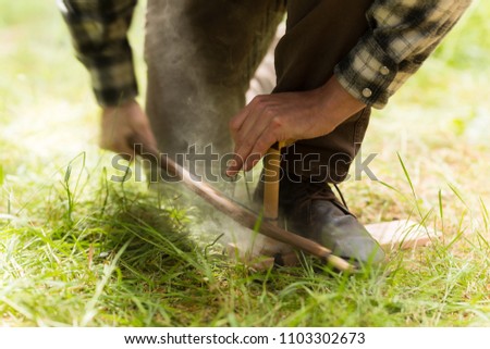 Man in plaid shirt using friction bow to start a fire in the forest Royalty-Free Stock Photo #1103302673