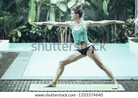 Content young woman standing on mat and doing yoga warrior two pose in swimming pool with blurred plants in background. Side view.