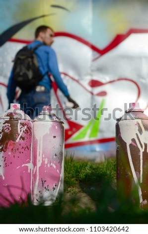 A few used paint cans against the background of the space with the wall on which the young guy draws a large graffiti drawing. Modern art of drawing walls in graffiti.