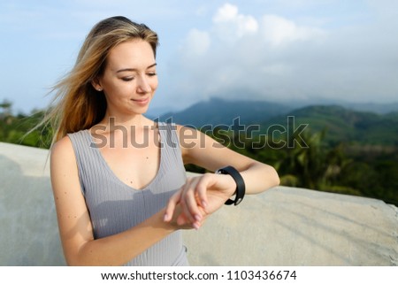 Young blonde woman looking at smart watch, green mouintains in background. Concept of modern etchnology and traveling, tourism and summer vacations.