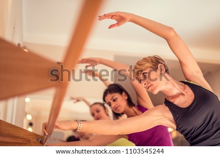 Beautiful adult women doing exercises on stretching ballet bar in Pilates class. Group of females doing yoga, pilates and fitness exercise indoors in studio. Royalty-Free Stock Photo #1103545244