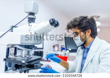 Lab technician is preparing to work. Scientist in lab coat and sterile mask doing microscope analysis. Life scientist researching in laboratory. Man Working in Laboratory With Microscope.
