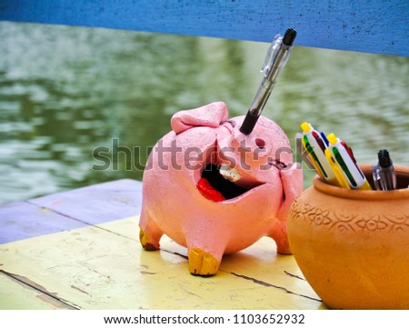Pink pig pottery pen holder on yellow wood table.Pen in pig nose  and smiles face with teeth.Soft focus water as background. Funny pen holder. Creative design for pen holder. Lovely stationery.