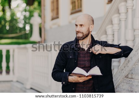 Horizontal portrait of attractive cheerful bald bearded male holds book, looks happily aside, enjoys calm atmosphere in royal park, reads something interesting, concentrated away. Reading concept