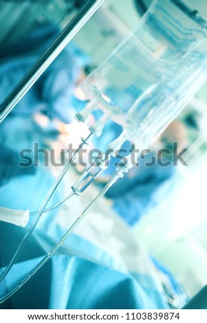 Hanging drip bag of saline on the background of working hard team of surgeons.