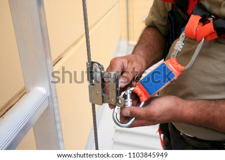 Clipping industrial Karabiner connected with fall arrest shock absorbing safety lanyard device into vertical lifeline steel cable system permanent that run through Aluminium safety ladder construction Royalty-Free Stock Photo #1103845949