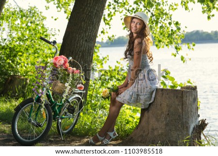 Beautiful Young Woman in a Dress and Hat on a Bicycle on the Nature in the Park Among the Grass and Trees