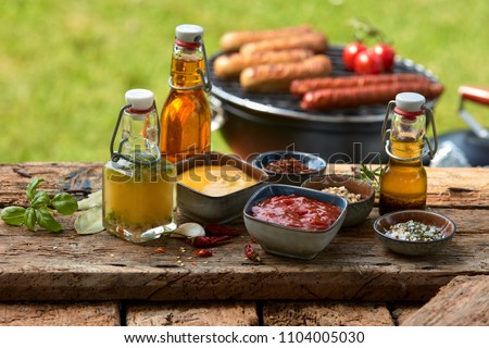 Assorted condiments and spices for a summer barbecue on a rustic wood picnic table outdoors in the garden with sausages grilling over a fire behind Royalty-Free Stock Photo #1104005030