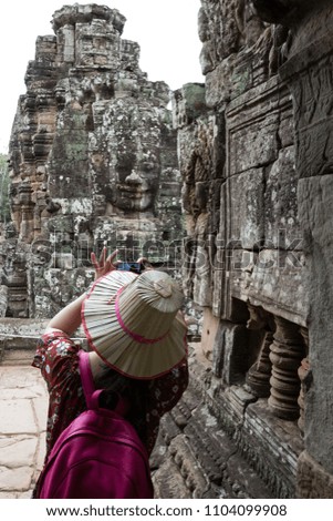 Woman traveler with pink backpack use her mobile phone taking a photo of ancient stone face of Bayon temple in Angkor Thom, Siem Reap province, Cambodia