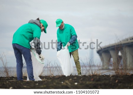 Two guys in greenpeace uniform picking up litter into big sacks while workin outdoors Royalty-Free Stock Photo #1104101072