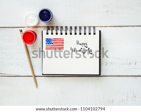 Notepad with a picture of the US flag and a handwritten inscription 4th July, a brush for drawing, colored gouache paints on a white, vintage wooden table. Top view, close-up