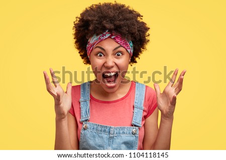 Portrait of annoyed African American female gestures and shouts in anger, quarrels with enemy, dressed in casual clothes, isolated over yellow studio background. Outraged dark skinned woman. Royalty-Free Stock Photo #1104118145