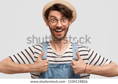 Portrait of happy delighted male gardener keeps thumbs raised, shows his like and approval, wears casual denim overalls and hat, smiles positively, isolated on white background. That`s fine!
