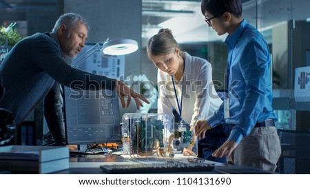 Team of Computer Engineers Lean on the Desk and Choose Printed Circuit Boards to Work with, Computer Shows Programming in Progress. In Background Technologically Advanced Scientific Research Center. Royalty-Free Stock Photo #1104131690