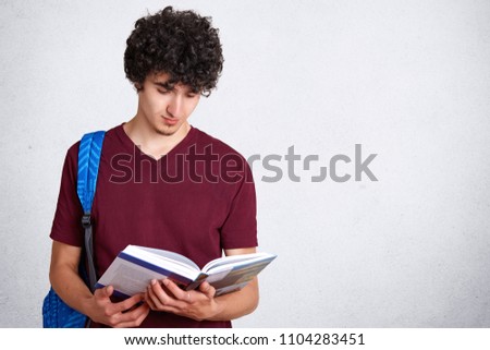 Concentrated male hipster reads attentively scientific literature, holds book, carries backpack, isolated over white background with blank copy space for your advertising content. Education concept