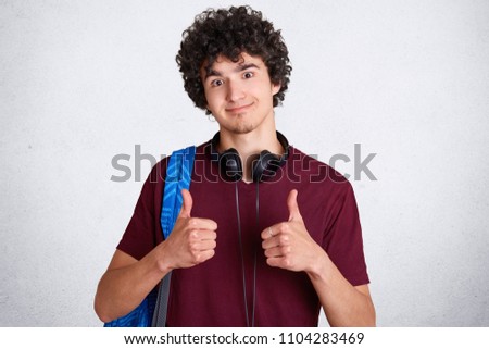 Attractive male student with crisp hair, wears casual t shirt, shows okay sign, carries rucksack, listens music via headphones, looks happily at camera, isolated on white background. People, education