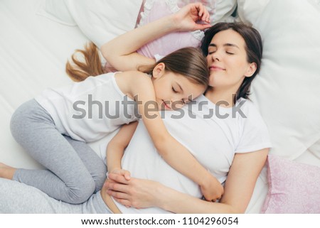 Pleased little child sleeps near her mother, embraces with love, has pleasant dreams, lie on comfortable bed. Mum and cute daughter have good sleep in bedroom. Family, sleeping and rest concept Royalty-Free Stock Photo #1104296054