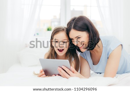 Positive cheerful small female kid and her young mother watch interesting cartoon on tablet, connected to wireless internet, pose against bedroom interior, have happy expressions. Family concept