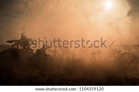 Medieval battle scene with cavalry and infantry. Silhouettes of figures as separate objects, fight between warriors on sunset foggy background. Selective focus Royalty-Free Stock Photo #1104359120