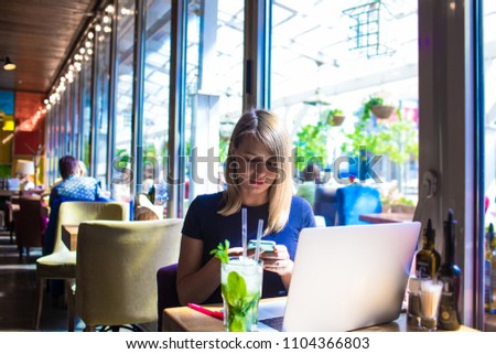 Young hipster girl chatting in social network via mobile phone, sitting with laptop computer and mojito beverage in modern restaurant interior. Woman skilled freelancer using apps on cellphone