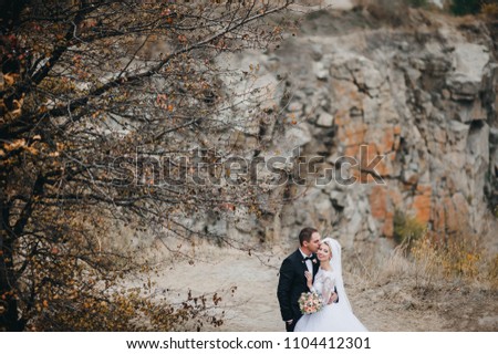 A stylish bridegroom in a black suit and a beautiful bride in a lace dress embrace and kiss against the background of rocks and river. Autumn wedding portrait of cute newlyweds.