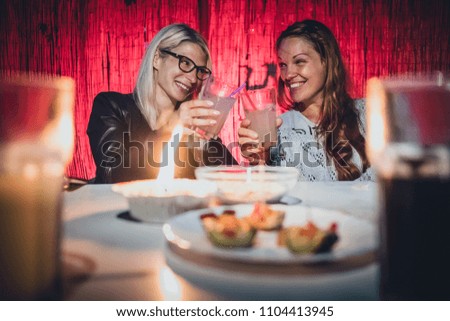 Two young women, a blonde and a redhead are cheering with glasses full of gin tonic drink and straws on a garden pary with candle on early evening