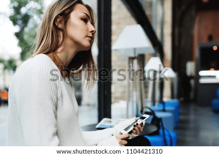 Portrait of charming blonde woman sitting beside a big window of a modern coffee shop with a mobile phone in her hands. Thoughtful student girl holding smartphone while sitting on a blurred background