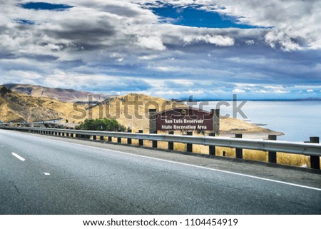 Driving through the golden hills of California; the San Luis Reservoir State Recreation area sign and dam on the right side of the road;