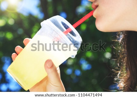 A cup of lemonade during a hot summer day in the hands of a girl, selective focus in top of dropped glass. The mouth of a girl who drinks at straw a cold lemonade in high summer temperatures.