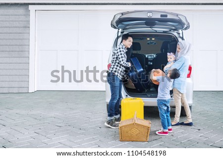 Picture of young father preparing suitcase into a car for holiday while standing with his family in the garage